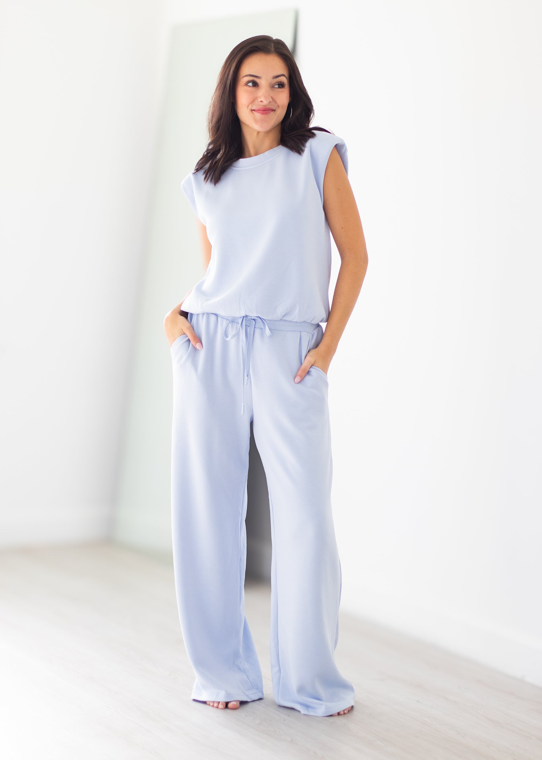 A woman modeling a light blue lounge set consisting of a sleeveless top and matching wide-leg pants, standing in a bright, minimalist room with white walls and light wood flooring.