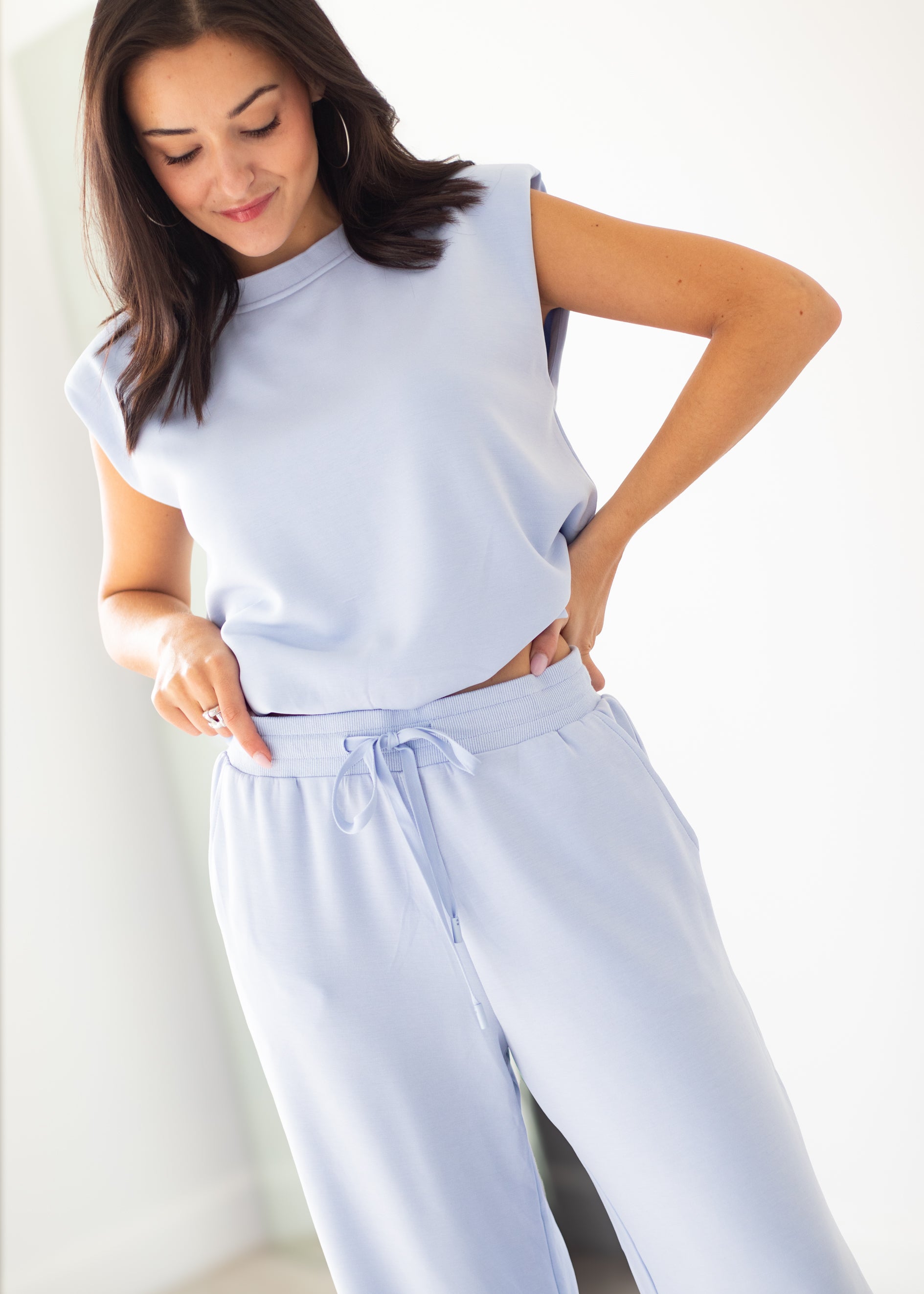 A woman modeling a light periwinkle blue two-piece lounge set consisting of a sleeveless mock-neck top and matching drawstring pants, set against a bright, neutral indoor background.