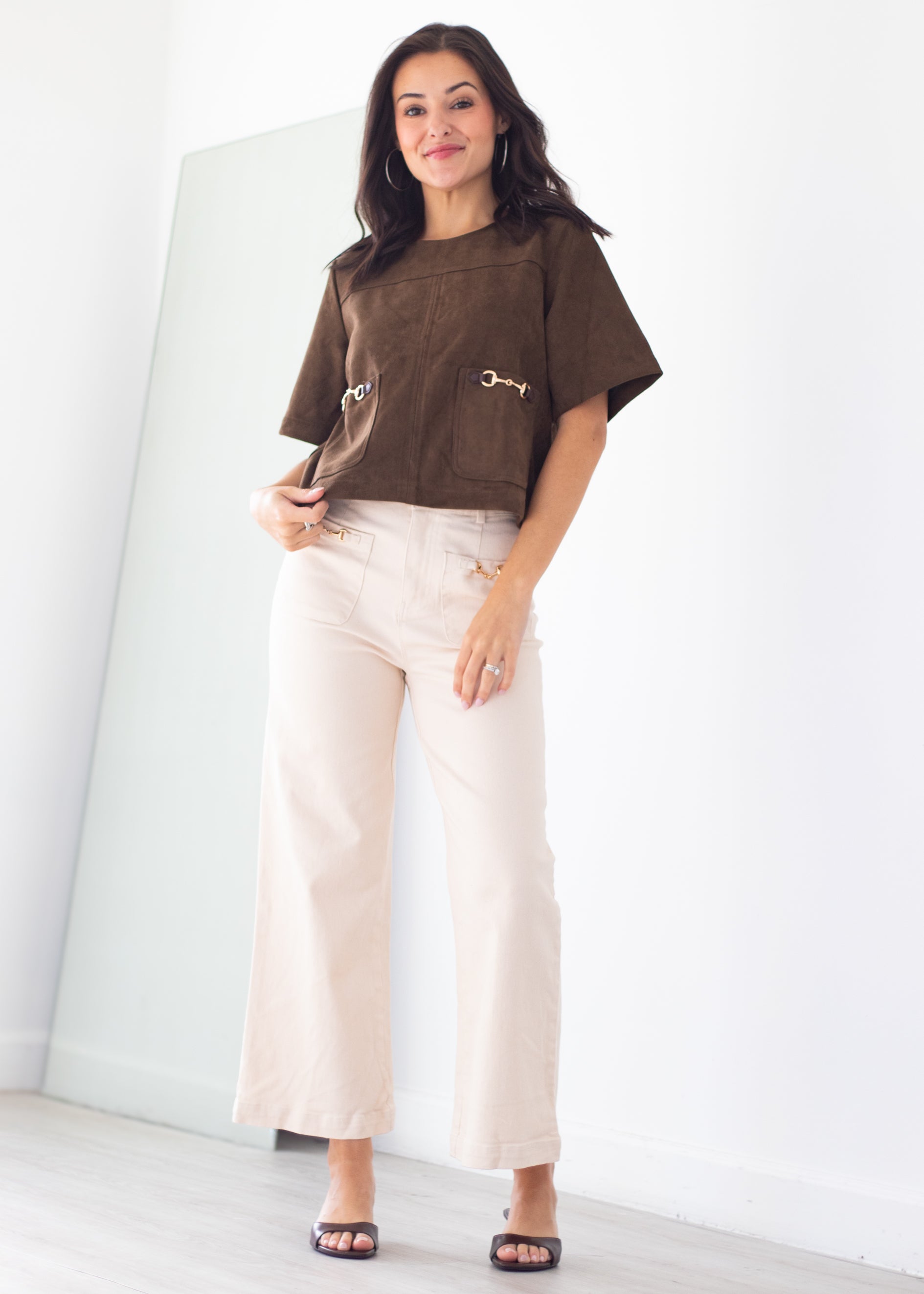 A woman modeling a brown short-sleeve cropped top with gold-tone hardware and cream wide-leg pants, standing in a bright indoor room with a large mirror on the left.