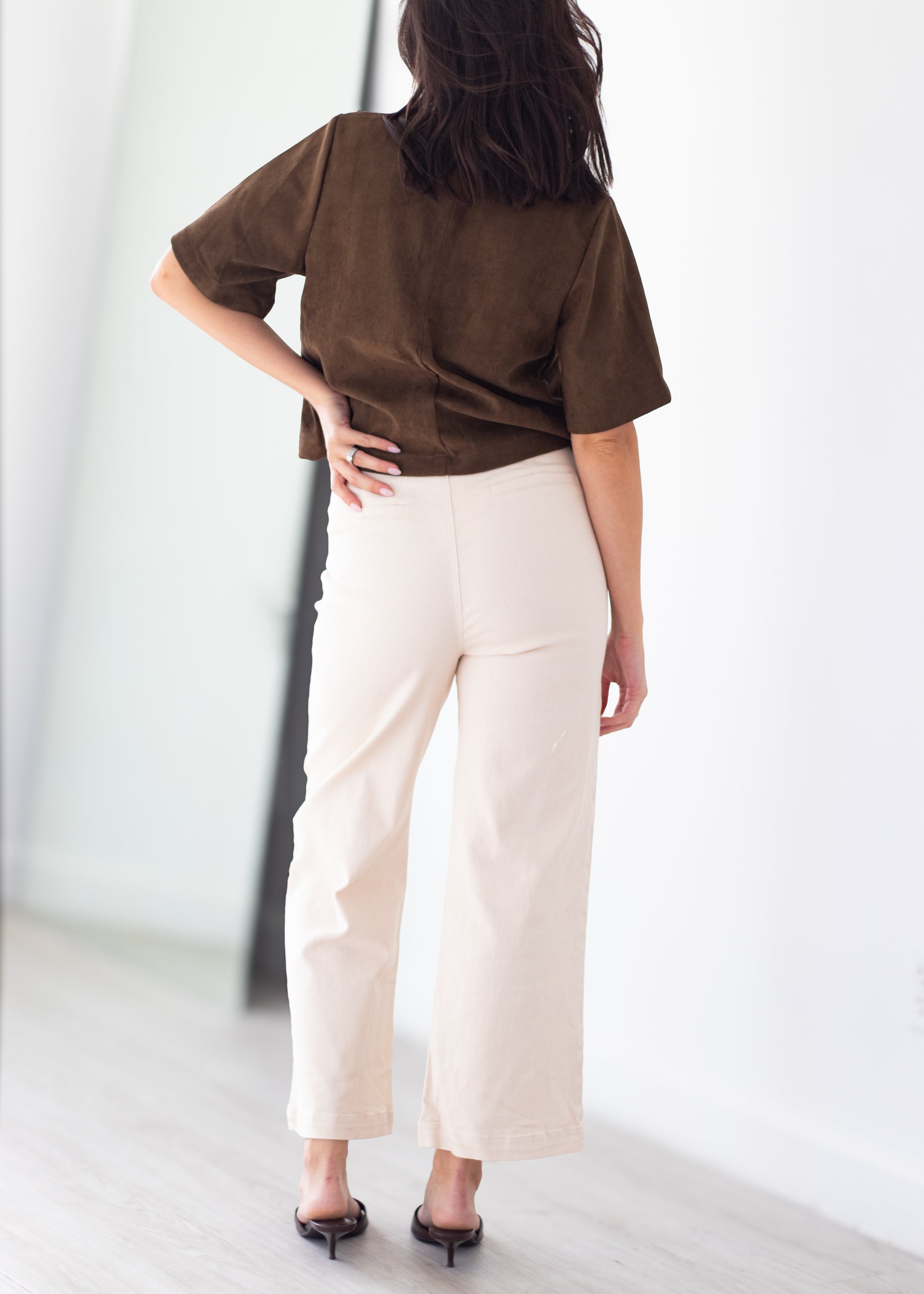 A woman shown from behind wearing a brown short-sleeve top and cream-colored wide-leg cropped pants, paired with black pointed-toe heels, in a bright minimalist room.