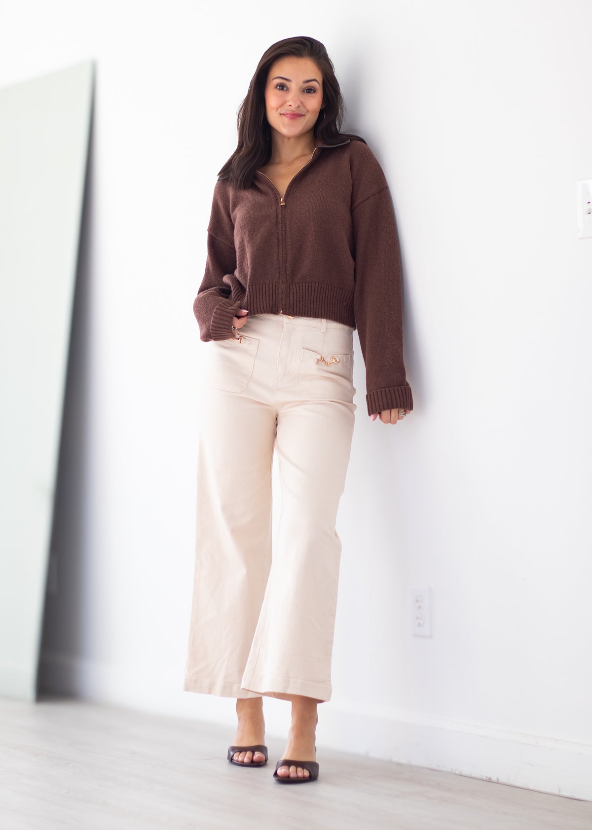 A woman stands against a white wall wearing a brown zip-up knit cardigan, cream wide-leg cropped pants, and black slide sandals.