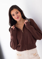 A woman wearing a brown ribbed knit zip-up sweater with a gray collar and cream pants, posing against a white studio background.