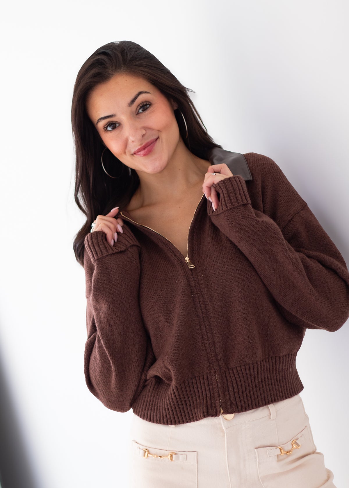 A woman wearing a brown ribbed knit zip-up sweater with a gray collar and cream pants, posing against a white studio background.