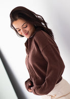 A woman wearing a brown ribbed knit zip-up sweater and cream pants, captured from a high-angle perspective against a bright white background.