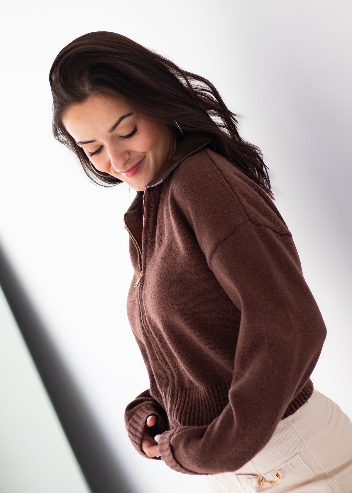 A woman wearing a brown ribbed knit zip-up sweater and cream pants, captured from a high-angle perspective against a bright white background.