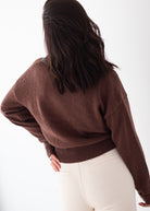 A woman shown from behind wearing a brown knit sweater and cream-colored pants against a plain white background.