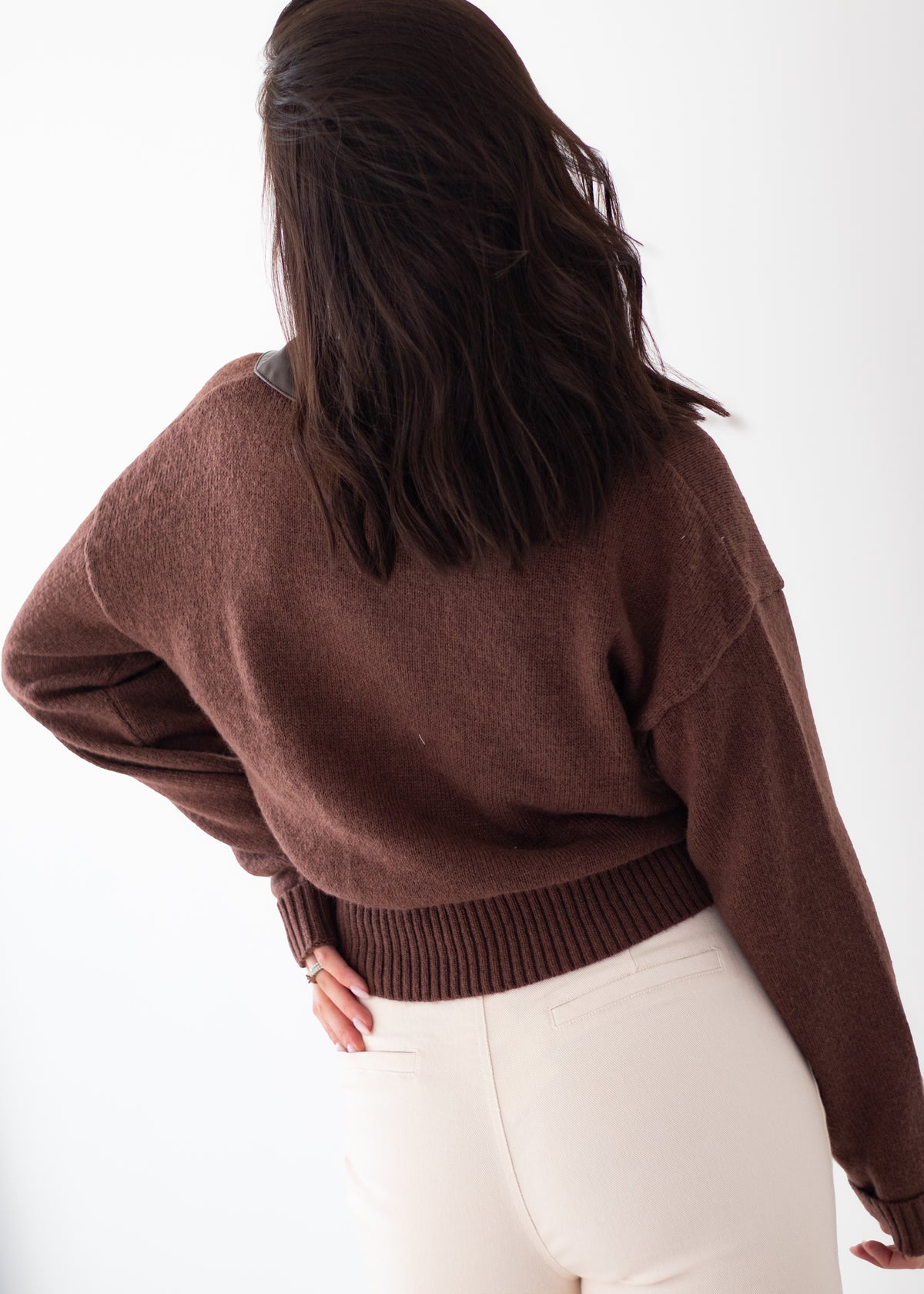 A woman shown from behind wearing a brown knit sweater and cream-colored pants against a plain white background.