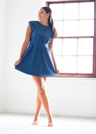 A woman modeling a sleeveless, flared blue dress in a bright room with a large window.