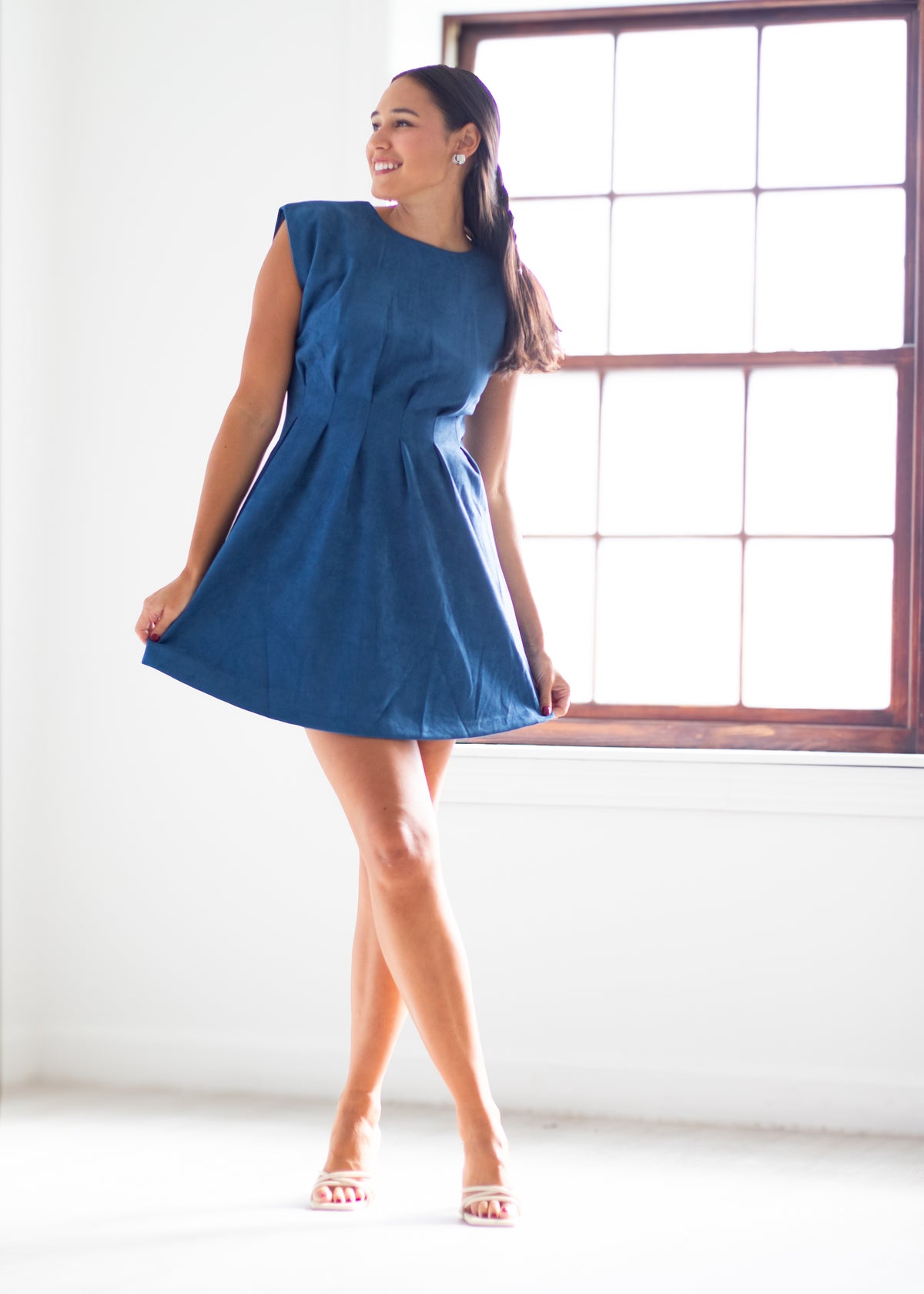 A woman modeling a sleeveless, flared blue dress in a bright room with a large window.