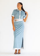 A woman modeling a light blue two-piece set consisting of a short-sleeve collared shirt with white trim and a matching high-waisted wrap skirt with a side slit, photographed against a plain white studio background.