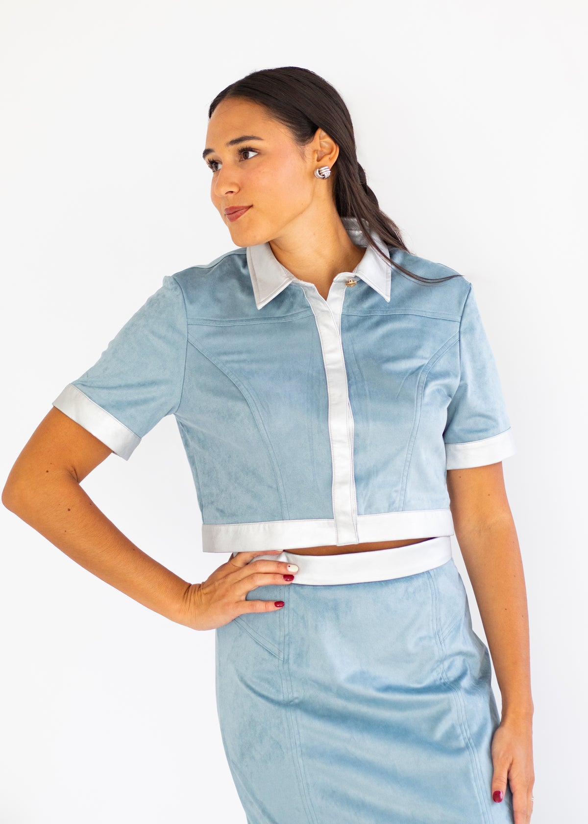 A woman modeling a light blue two-piece outfit consisting of a short-sleeve collared shirt with white trim and a matching light blue skirt with a white waistband, against a plain white background.