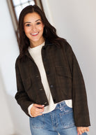 A woman wearing a dark brown cropped plaid jacket over a cream ribbed knit sweater and blue jeans, standing in a brightly lit indoor setting.