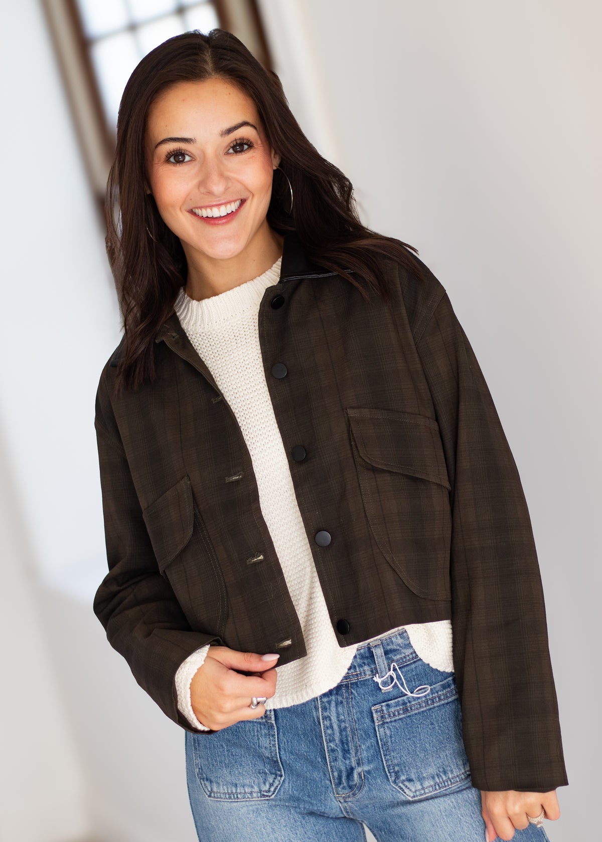 A woman wearing a dark brown cropped plaid jacket over a cream ribbed knit sweater and blue jeans, standing in a brightly lit indoor setting.