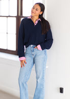 A woman wearing a navy long-sleeve polo-style sweater with pink contrast collar, placket, and cuffs, paired with light-wash blue jeans, standing in a bright indoor setting.
