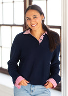 A woman smiling and wearing a navy blue knit polo sweater with pink and white striped collars and cuffs, paired with light blue jeans, standing in front of a bright window.