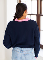 A woman viewed from behind wearing a navy blue knit sweater with a pink and white striped collar, paired with light blue jeans in a bright indoor setting.
