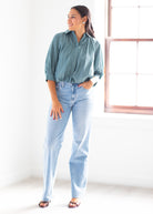 A woman modeling a sage green button-down shirt and light-wash blue jeans in a bright, minimalist room with a window.