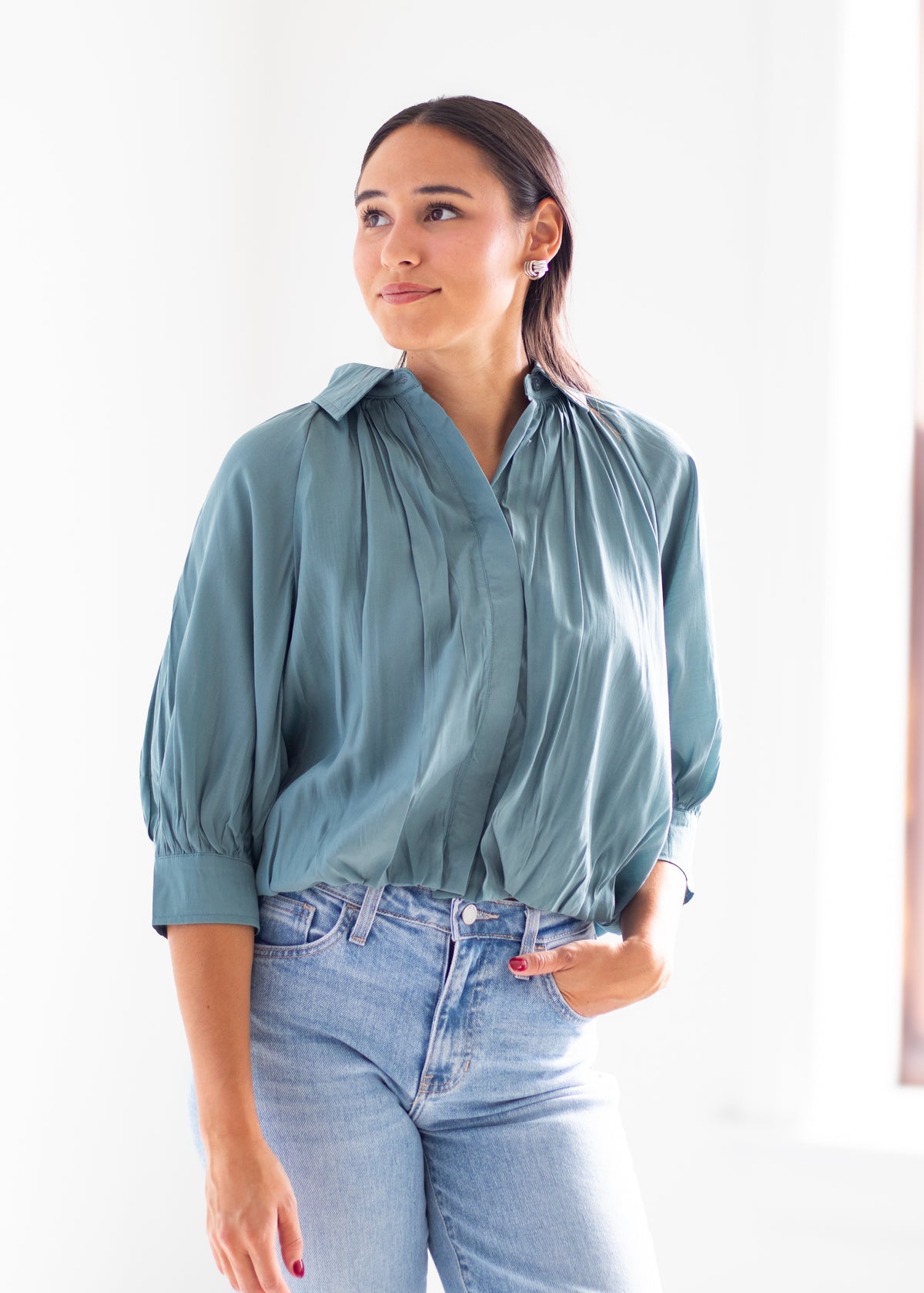 A woman wearing a dusty teal blue blouse with voluminous elbow-length sleeves and a gathered neckline, paired with light-wash blue jeans, standing against a bright white background.