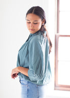 A woman wearing a dusty blue high-neck blouse with three-quarter sleeves and light-wash blue jeans, standing in a brightly lit room next to a window.