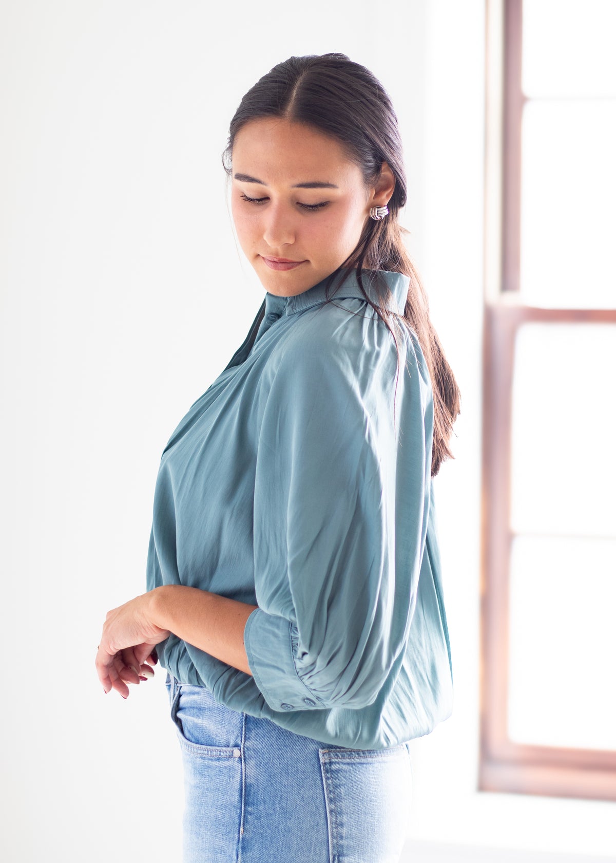 A woman wearing a dusty blue high-neck blouse with three-quarter sleeves and light-wash blue jeans, standing in a brightly lit room next to a window.