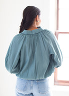 A woman viewed from behind wearing a dusty blue long-sleeve blouse with a gathered, pleated back and light-wash blue jeans, standing in a brightly lit room.