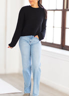 A woman wearing a black ribbed knit sweater and light-wash blue jeans, standing in a bright indoor room with a large window.