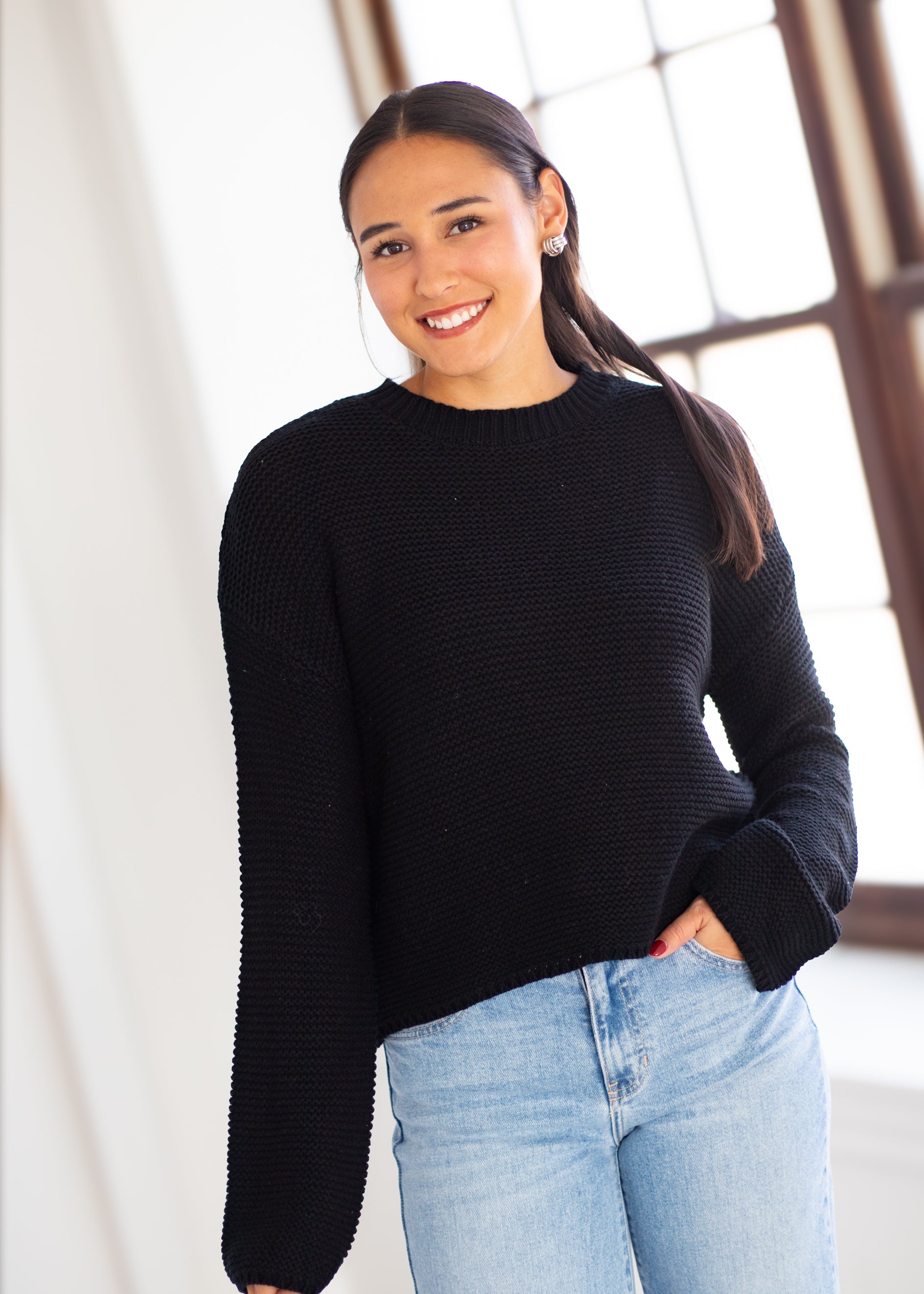 A woman wearing a black ribbed knit long-sleeve sweater and light blue jeans, standing in a bright indoor setting with a large window in the background.