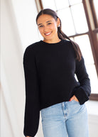 A woman wearing a black ribbed knit long-sleeve sweater and light blue jeans, standing in a bright indoor setting with a large window in the background.