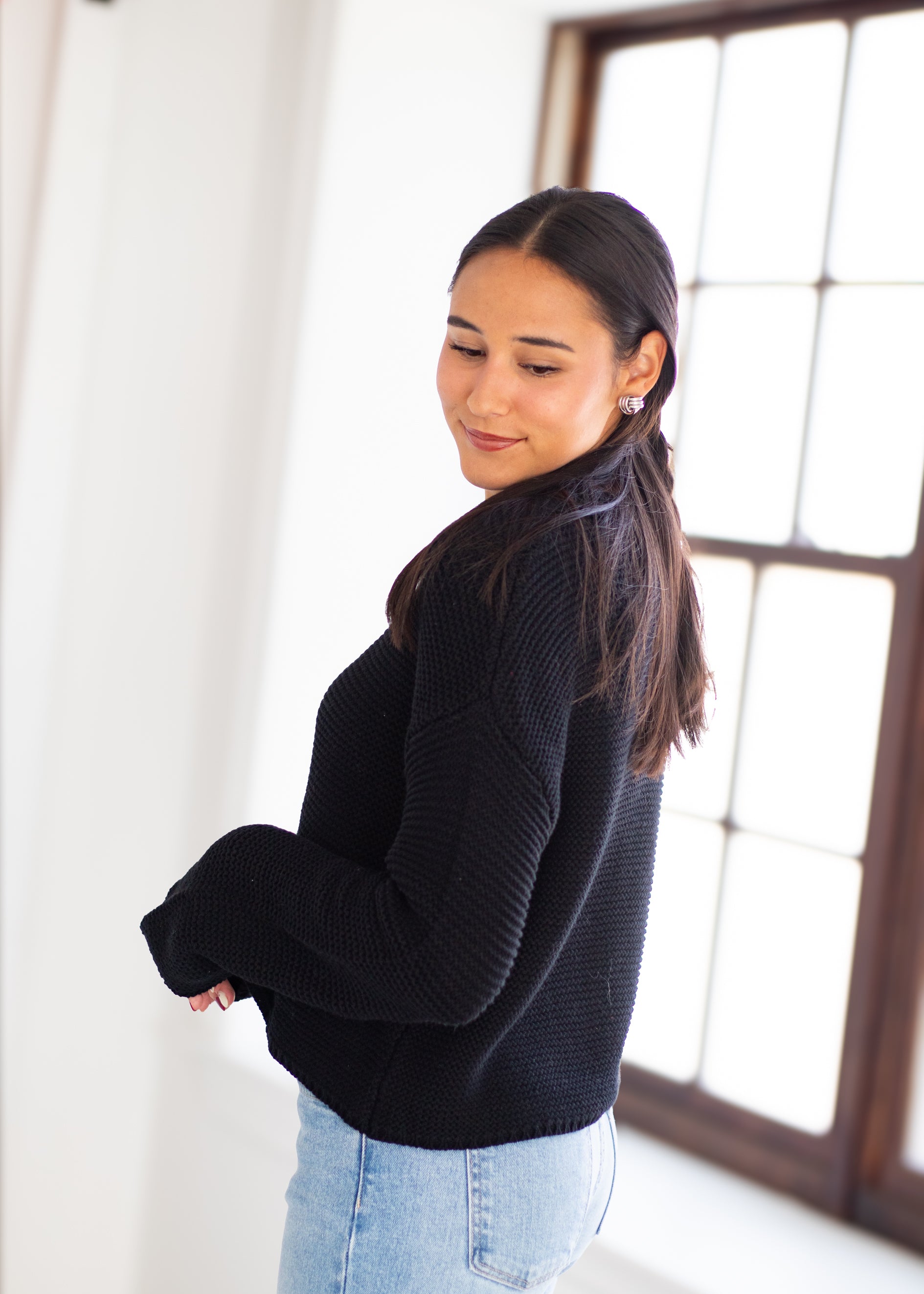A woman wearing a black ribbed knit sweater and light blue jeans, standing in a bright room with a large window in the background.