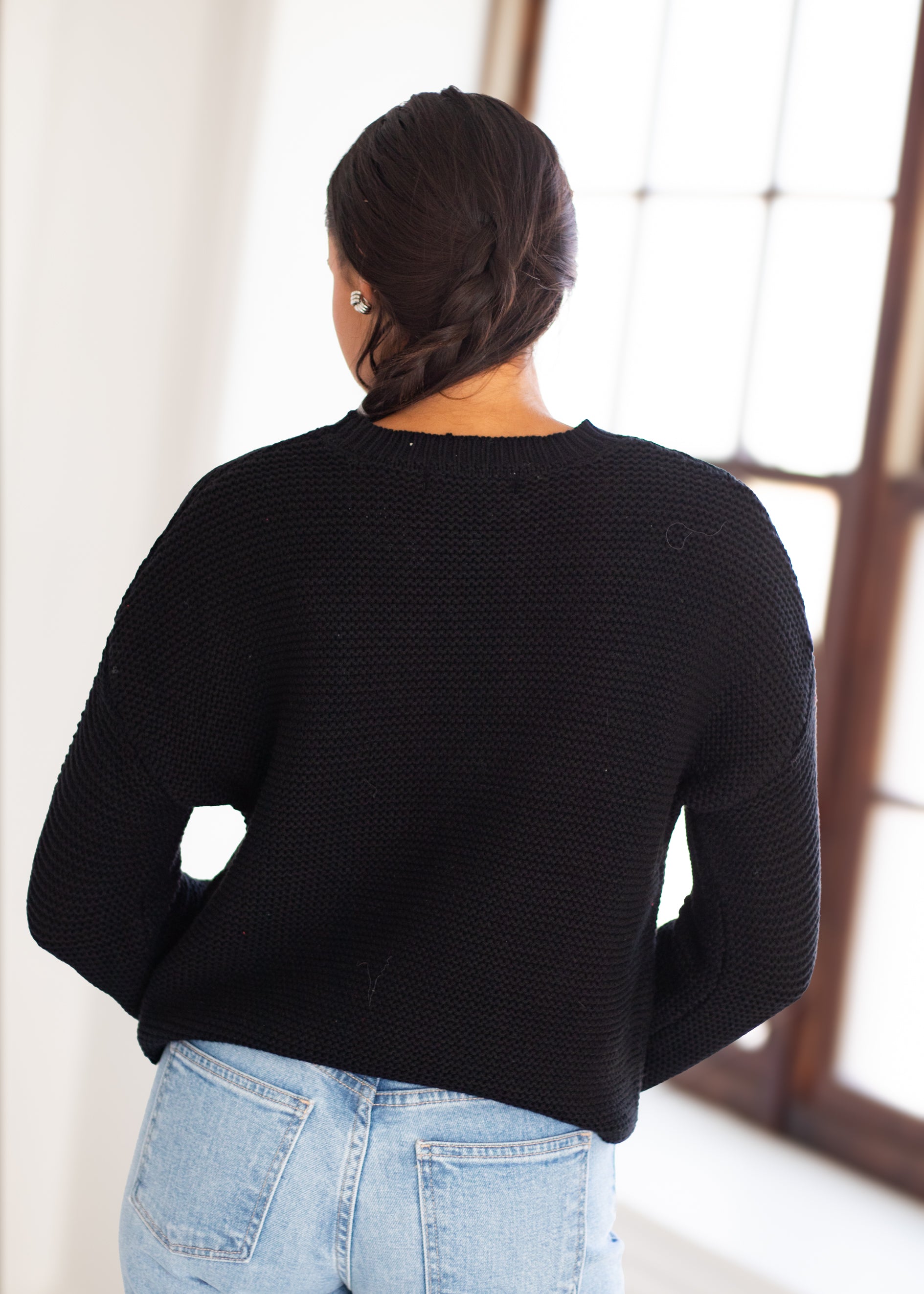 A woman viewed from behind wearing a black textured knit sweater and light blue jeans, standing in a bright indoor setting near a window.