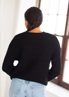 A woman viewed from behind wearing a black textured knit sweater and light blue jeans, standing in a bright indoor setting near a window.
