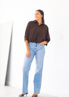 A woman modeling a dark brown pleated button-down shirt and light-wash blue jeans, standing against a bright white wall.