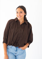 A woman wearing a dark brown, gathered long-sleeve blouse tucked into light wash blue jeans, standing against a solid white background.