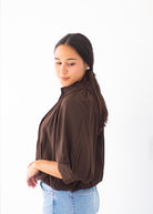 A woman with dark hair tied back is wearing a dark brown long-sleeve blouse with gathered shoulders and light blue jeans, standing against a plain white background.