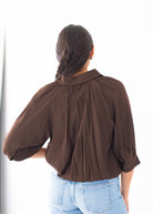 A woman viewed from behind wearing a dark brown long-sleeve blouse with voluminous puff sleeves and a gathered neckline, paired with light blue jeans against a bright white background.