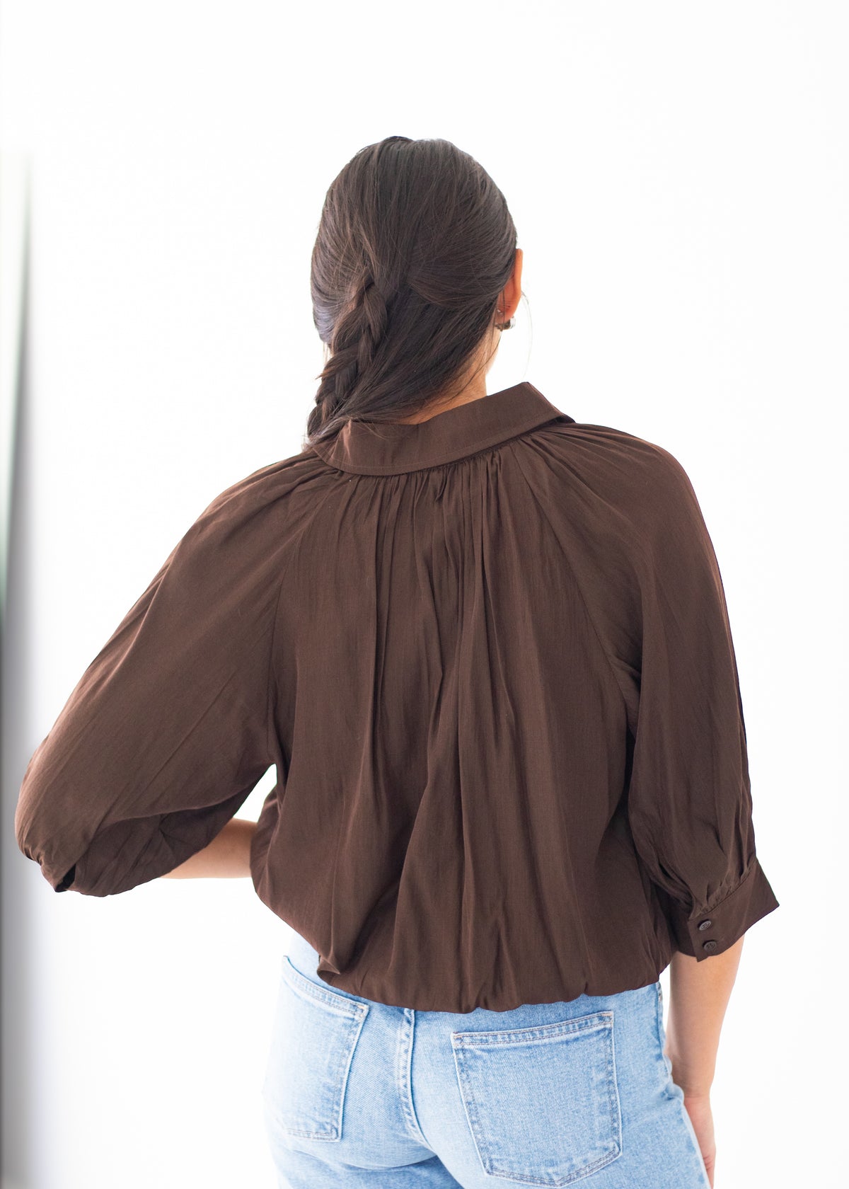 A woman viewed from behind wearing a dark brown long-sleeve blouse with voluminous puff sleeves and a gathered neckline, paired with light blue jeans against a bright white background.