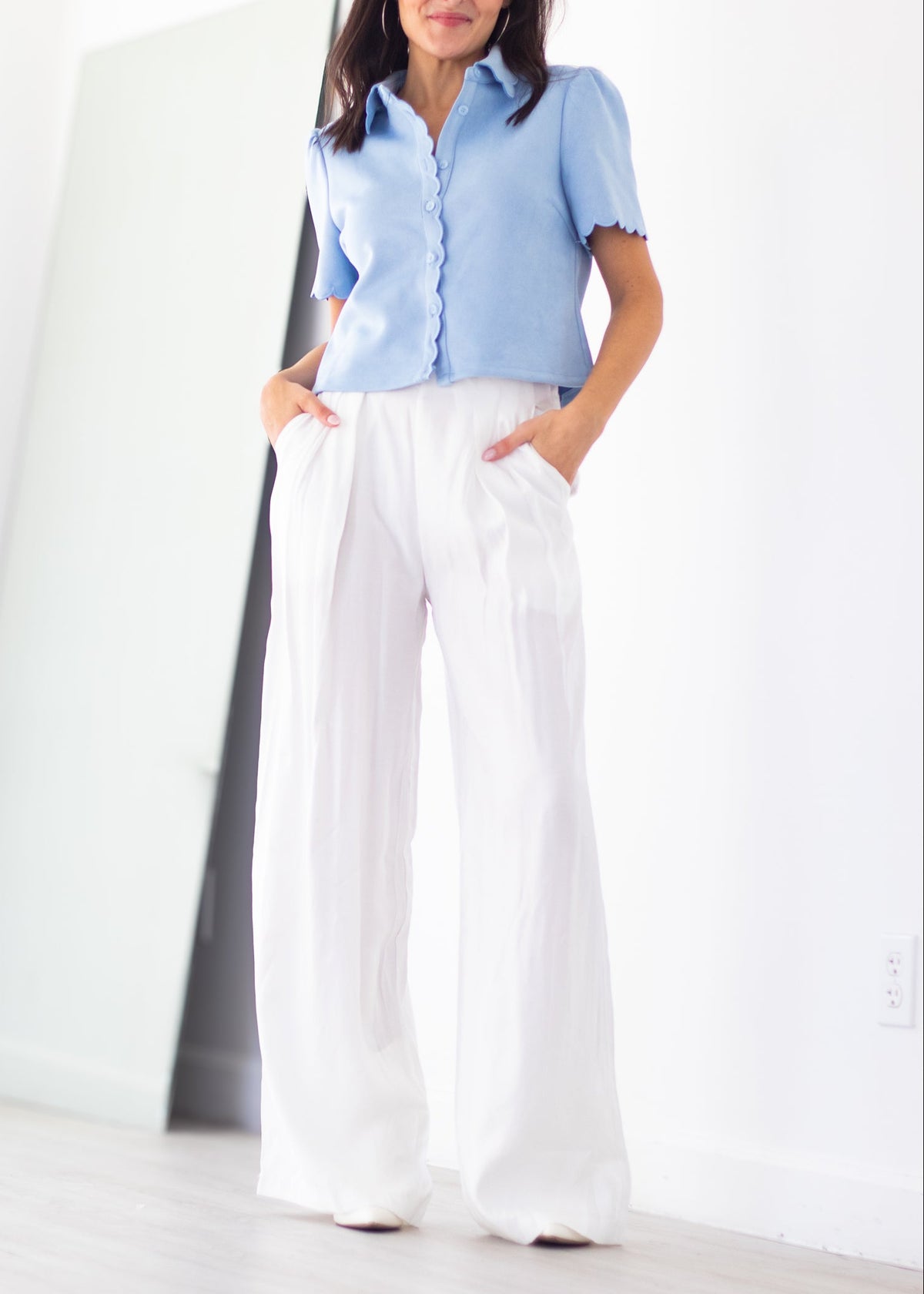 A woman modeling a light blue short-sleeve button-up shirt with scalloped edges and white wide-leg pants, standing in a bright, minimalist room with a large mirror against a white wall.