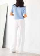 A woman viewed from behind wearing a light blue short-sleeve top and white wide-leg pants, standing in a bright indoor room with a mirror and light wood floor.