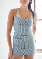 A woman wearing a green and white gingham mini dress with spaghetti straps, a sweetheart neckline, and a matching fabric belt with a decorative buckle, shown in a brightly lit indoor setting.