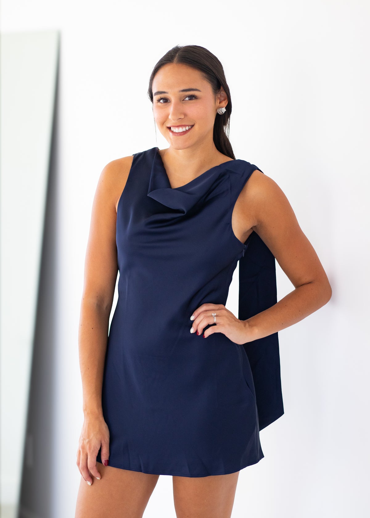 A woman modeling a sleeveless navy blue cowl-neck mini dress with side pockets, standing against a plain white wall.