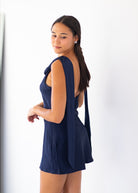 A woman modeling a navy blue sleeveless mini dress with a draped back detail, shown from a side-back angle against a plain white studio background.