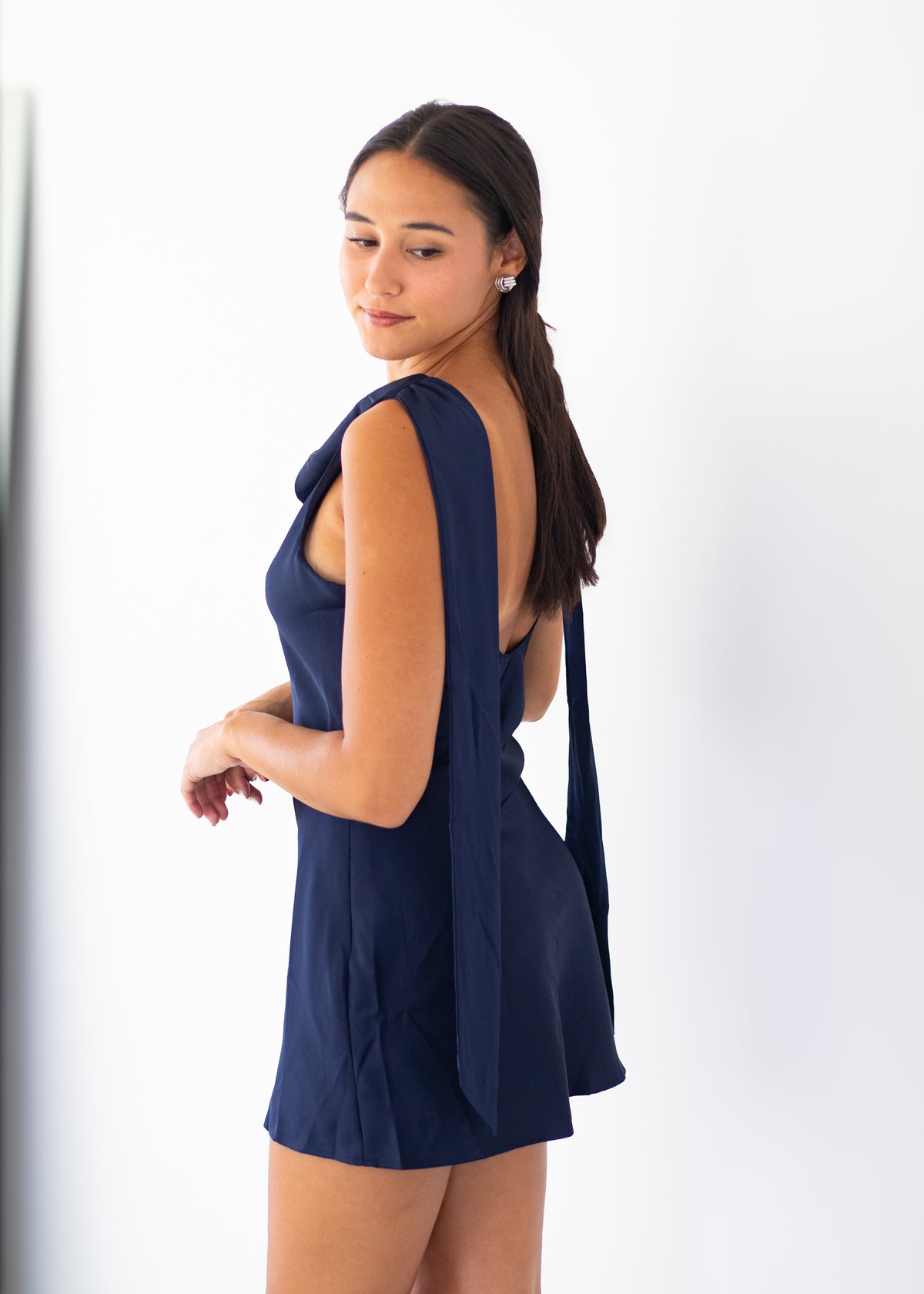 A woman modeling a navy blue sleeveless mini dress with a draped back detail, shown from a side-back angle against a plain white studio background.