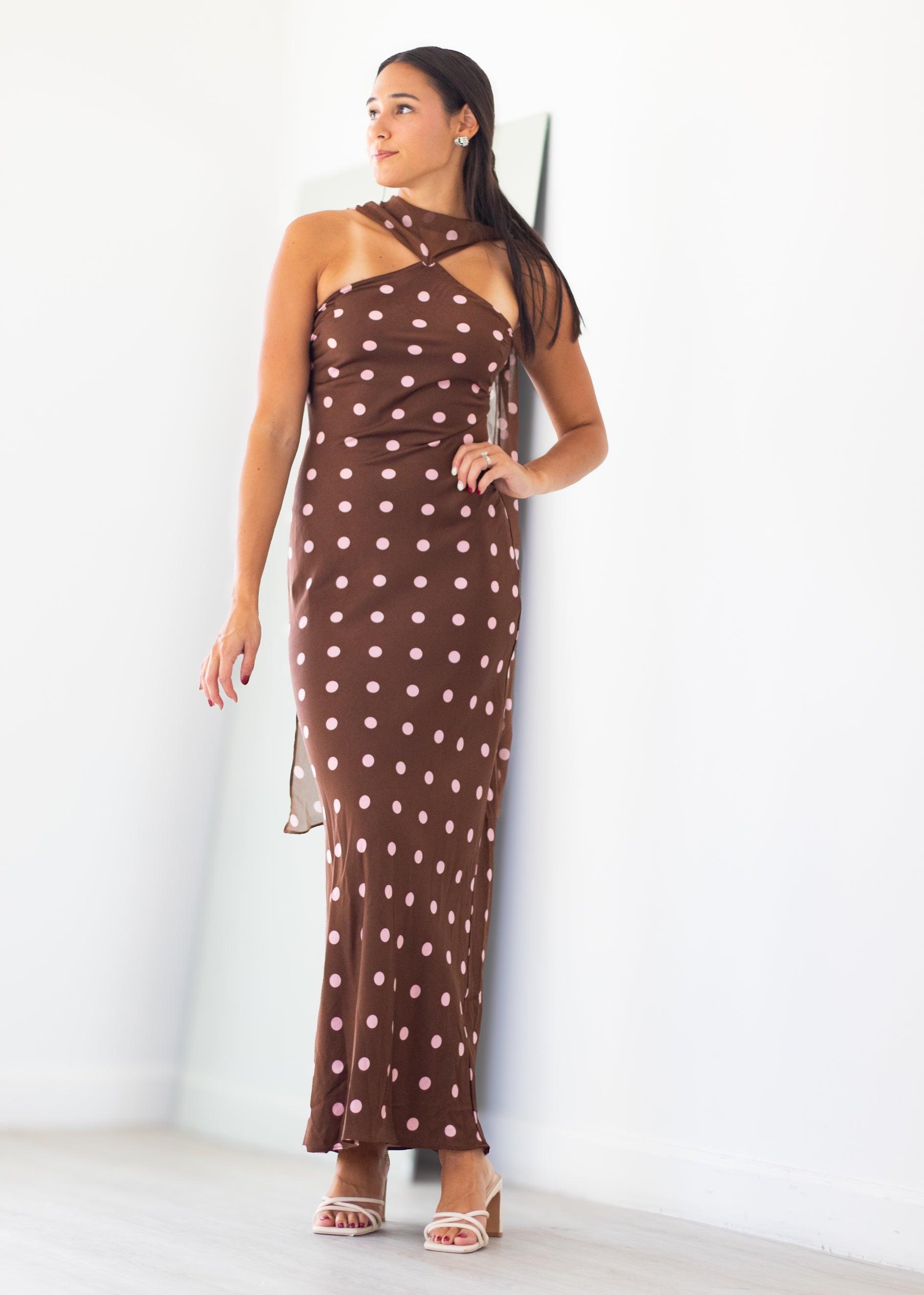 A woman modeling a brown halter-neck maxi dress with a white polka dot pattern, standing against a plain white wall.