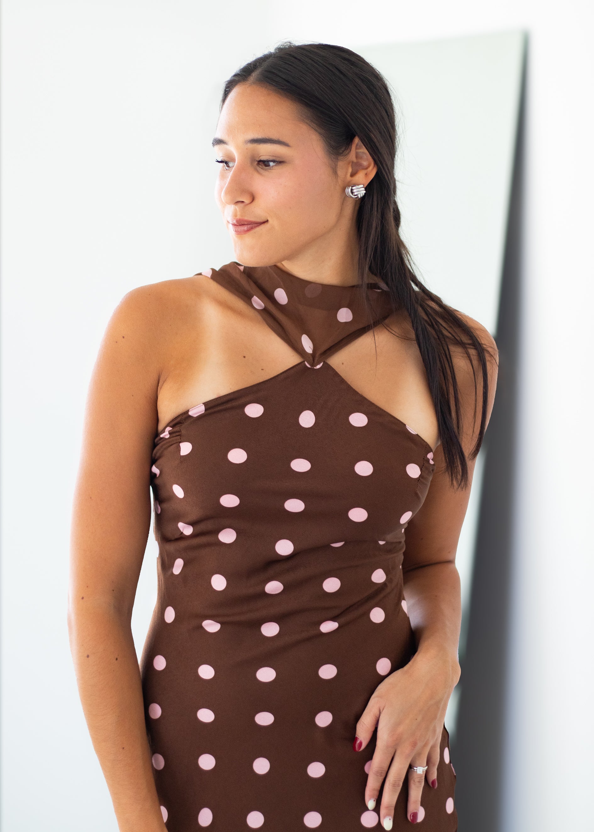 A woman wearing a brown halter-neck dress with light pink polka dots, standing against a white wall.