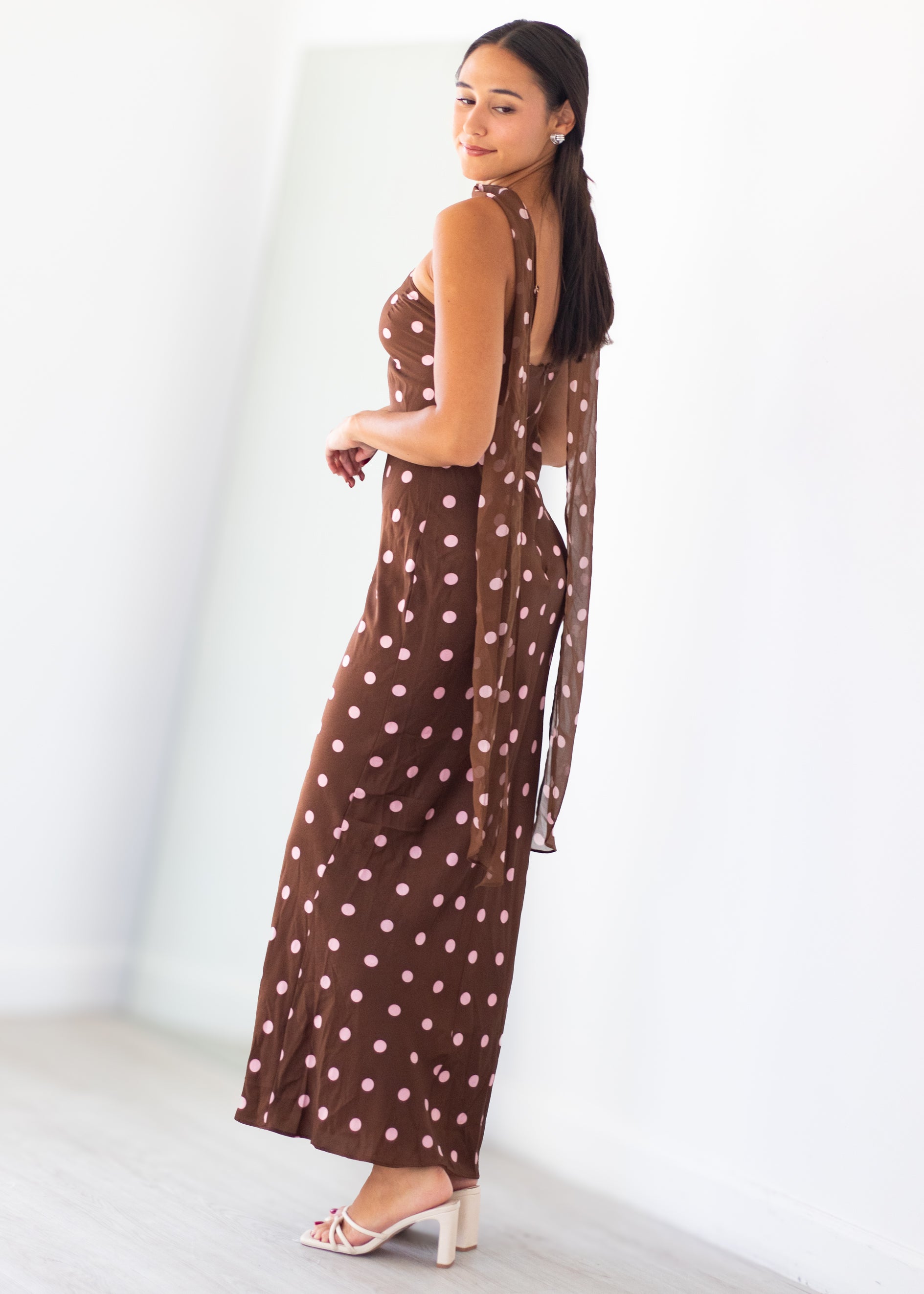 A woman modeling a brown halter-neck maxi dress with pink polka dots, shown from a side-back angle in a bright, minimalist studio setting.