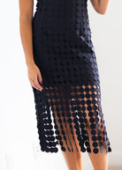 A close-up of a model wearing a black midi dress covered in overlapping circular cutout embellishments, with a fringe effect at the hem, against a plain white background.