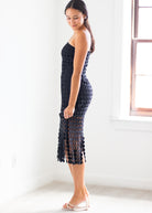 A woman modeling a black crochet knit strapless midi dress with a scalloped edge and openwork pattern, standing in a bright white room next to a window.