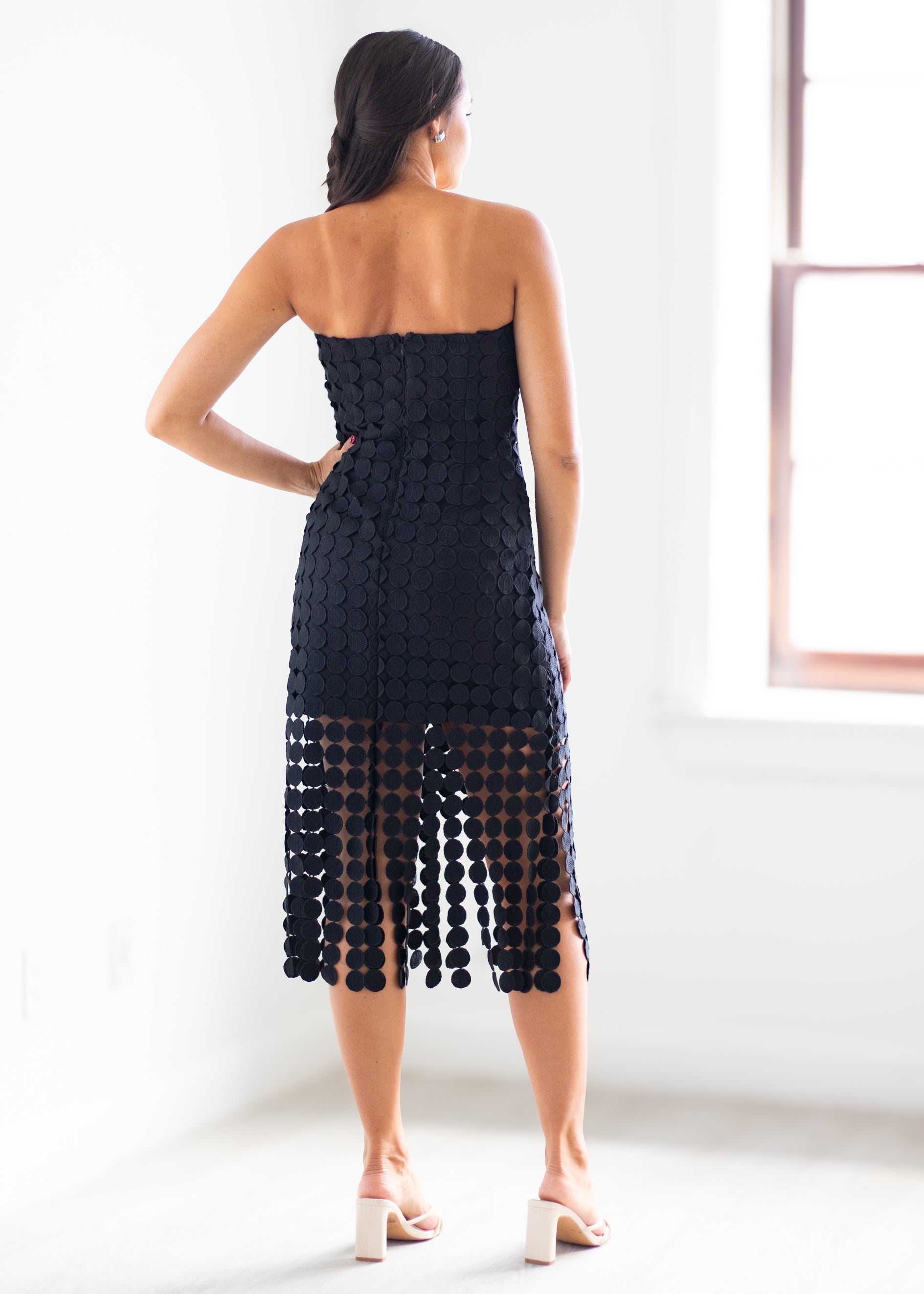 A woman shown from the back wearing a strapless black crochet midi dress with a textured pattern and a sheer lower section with hanging crochet details, paired with white block-heel sandals in a bright white room.