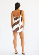 A woman viewed from behind wearing a brown and white diagonally striped mini dress with an open back and tie detail, paired with black pointed-toe heels, against a plain white studio background.