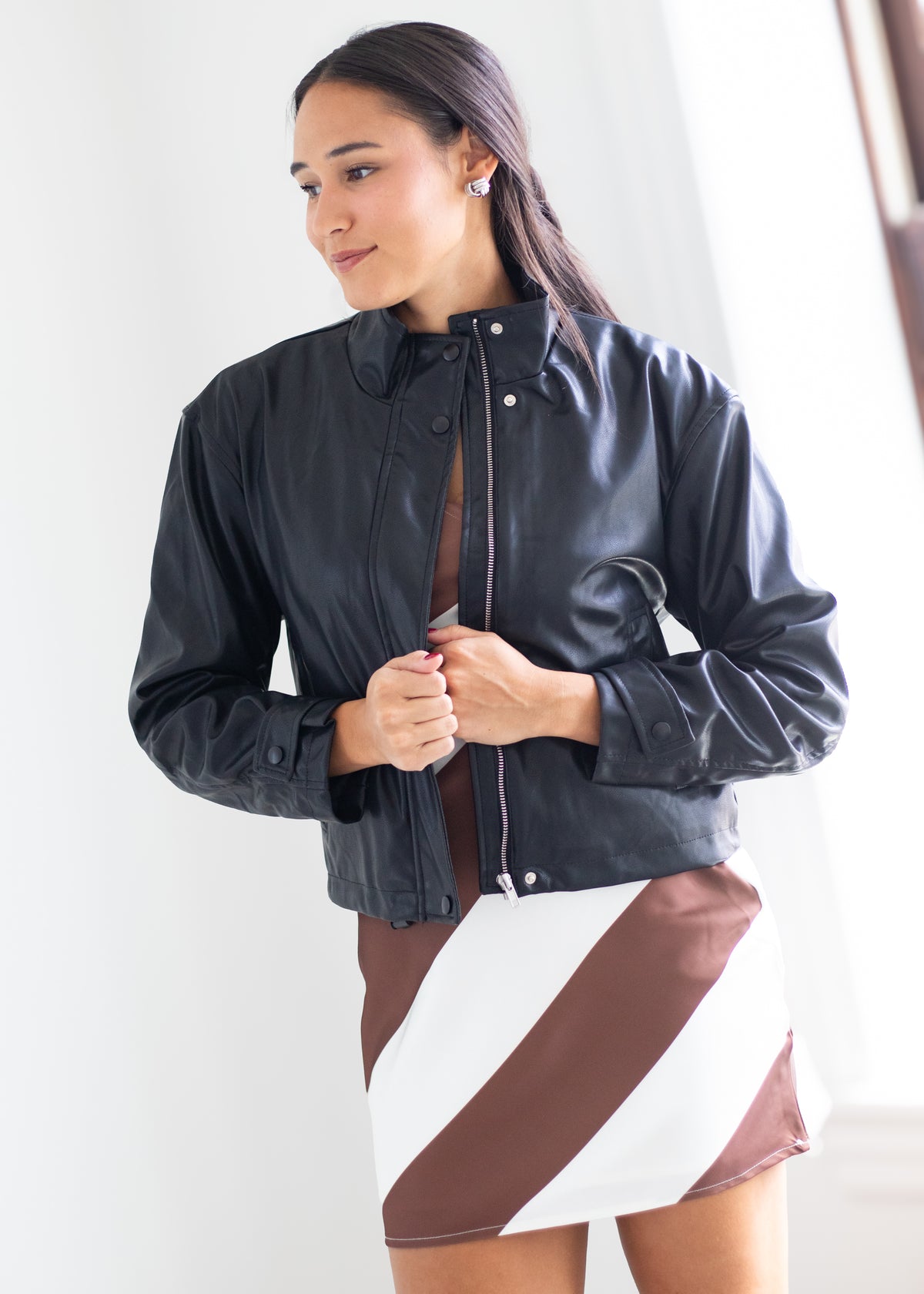 A woman wearing a black faux-leather cropped jacket over a brown top and a brown, white, and gray striped skirt, standing against a bright white background.